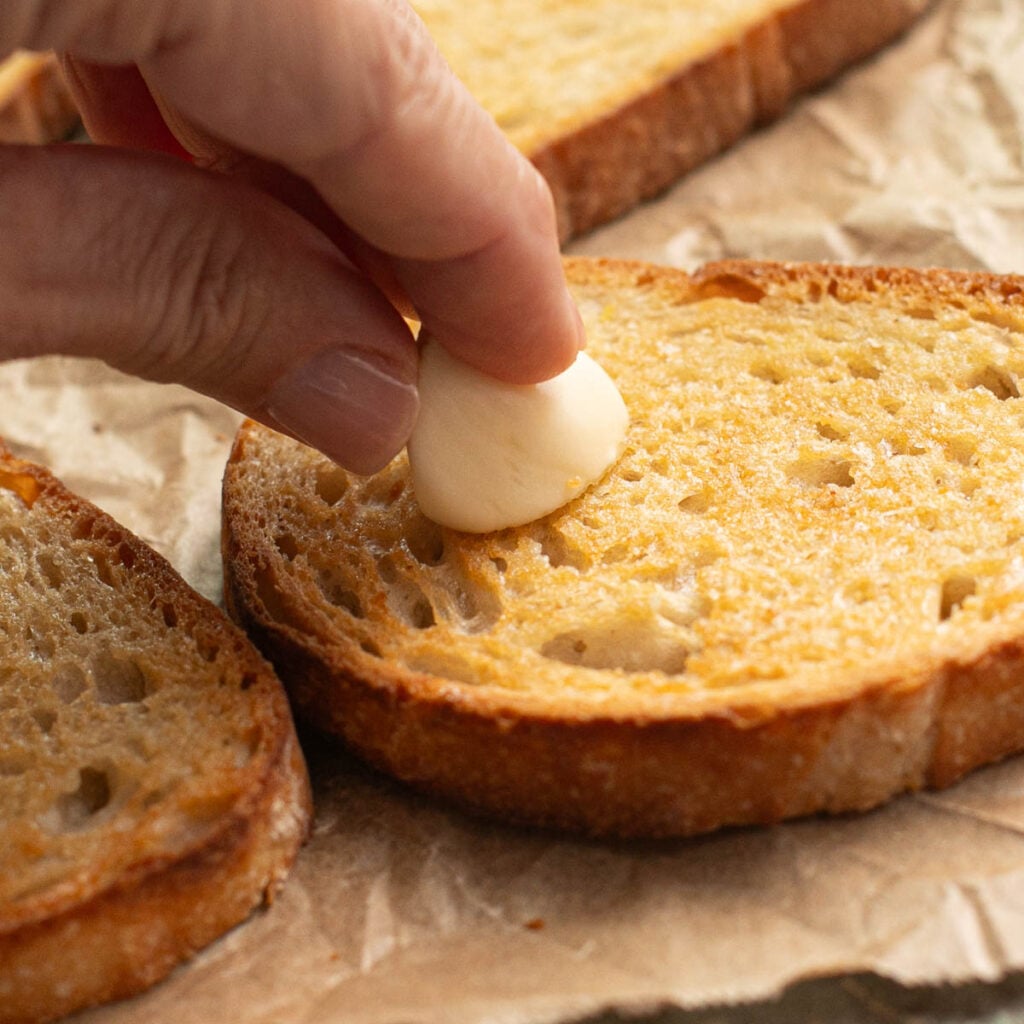 a hand rubbing a garlic clove onto sourdough toast