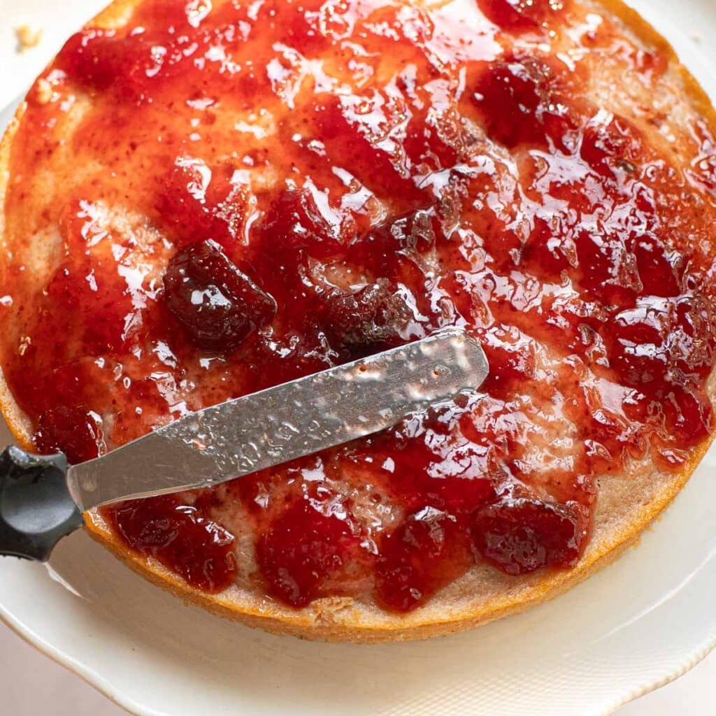 a spatula spreading strawberry jam on a layer of cake