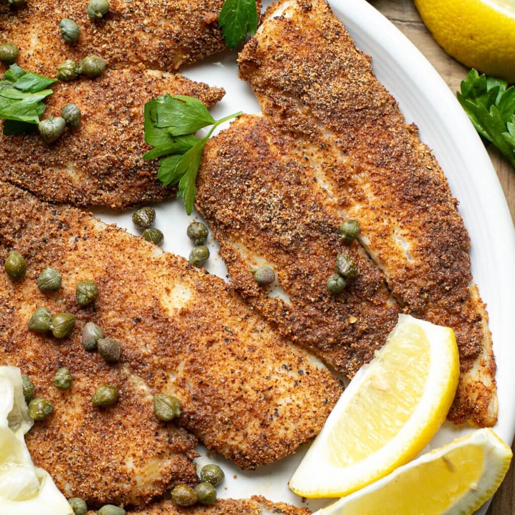 air fryer flounder on a white plate topped with fresh parsley, capers and lemon wedges