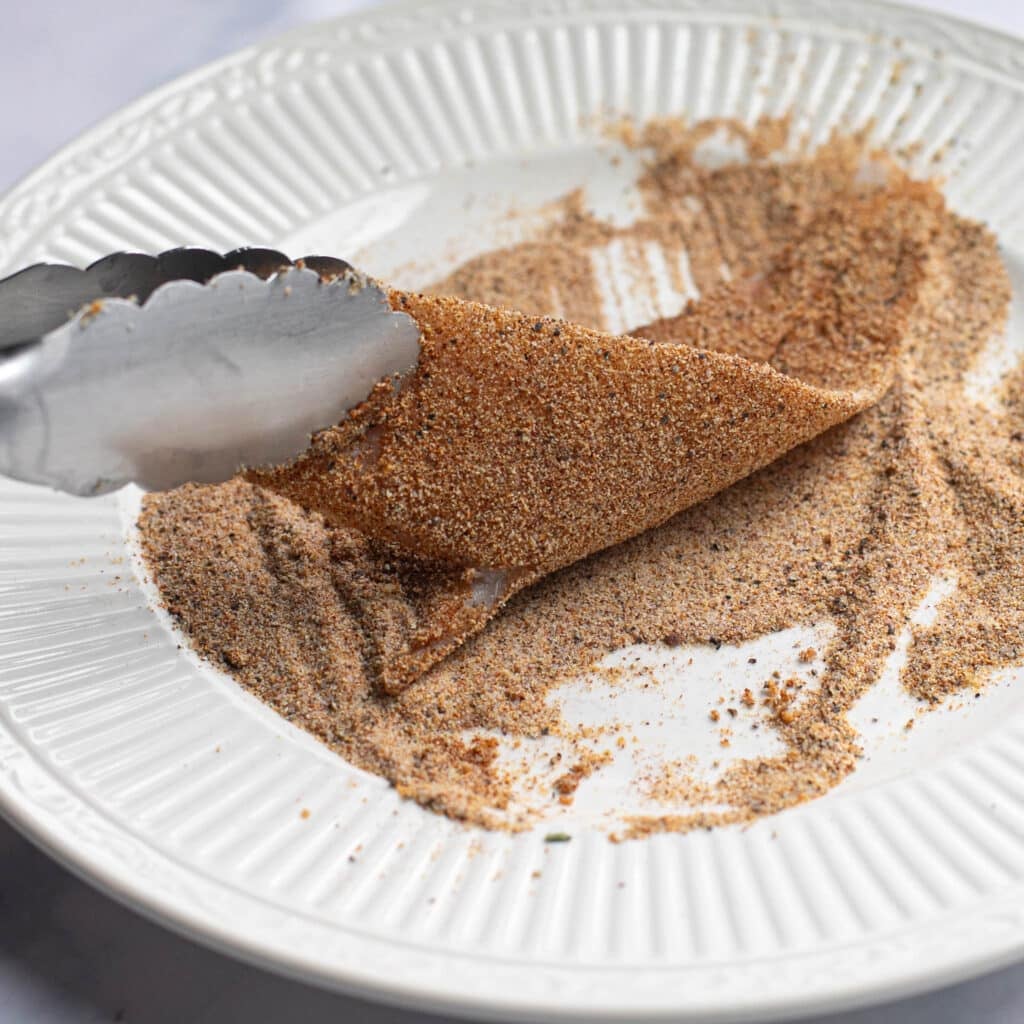 metal tongs holding a piece of flounder while it is coated in dry spices