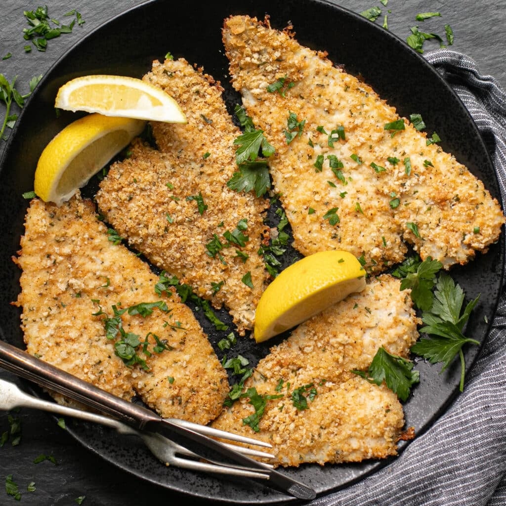 Parmesan panko flounder fillets on a black plate garnished with lemon wedges and fresh parsley