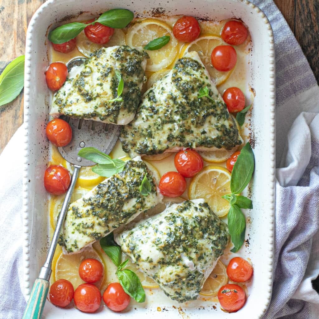 baked snapper in a casserole dish with pesto sauce, burst tomatoes and lemons