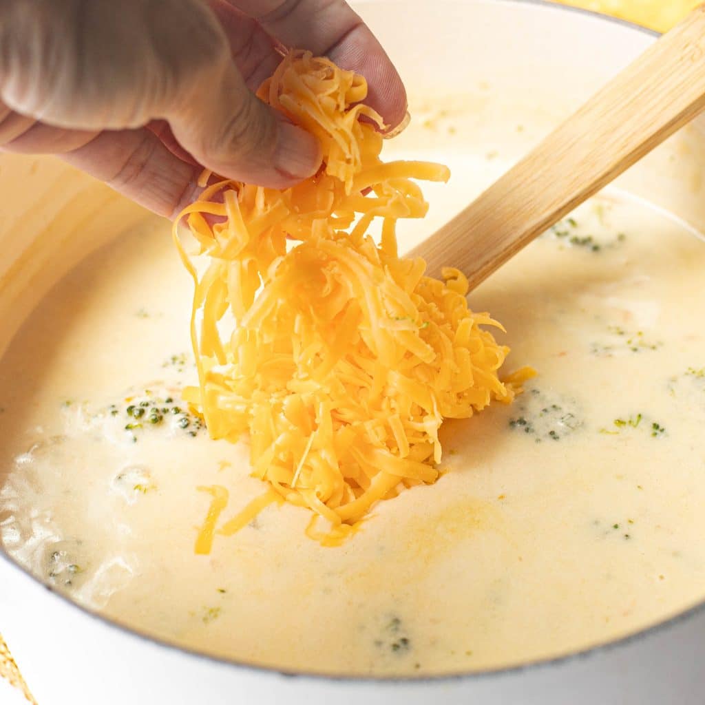a hand putting shredded cheddar cheese into broccoli soup