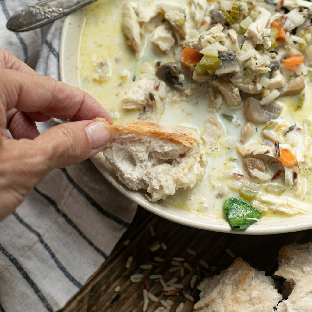 a hand dipping bread into a bowl of chicken and wild rice soup
